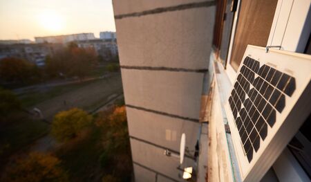 Solar panel on the window of a high-rise residential building against a background of a sleeping area of the city with clear blue sky on a warm day. Converting solar radiation into electricityの写真素材