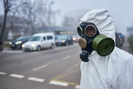Selective focus of scientist wearing gas mask and protective costume looking at road with cars, waiting for green light. Close up of man posing near pedestrian crossing. Environmental disaster, fog.の写真素材