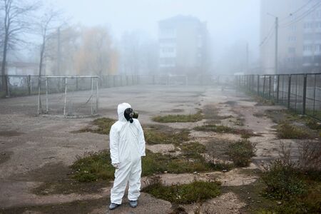 From above view of scientist looking aside. Man in gas mask and protective costume standing on empty football pitch in city yard near buildings. Environmental disaster, smoky atmosphere, fog.の写真素材