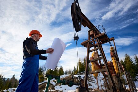 Petroleum engineer in blue uniform and orange helmet standing in oil field with printed plan next to the oil pump jack, on beautiful sunny day. Concept of petroleum industry and oil extraction.の写真素材