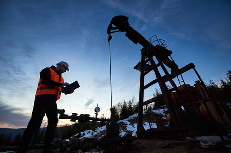 Oil engineer in work vest at oil pump rocker-machine, making notes while checking work of balanced...