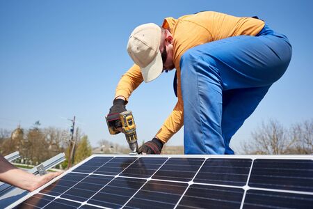 Male engineer installing stand-alone solar photovoltaic panel system using screwdriver. Electrician mounting blue solar module on roof of modern house. Alternative energy ecological concept.の写真素材