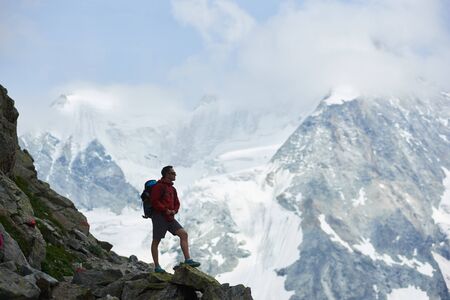 Side view of tourist with backpack standing on big stone, looking at beautiful mountains with snow....