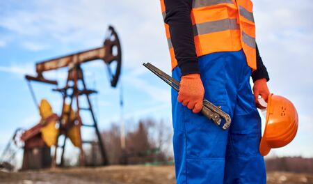 Close up of petroleum operator in work orange vest holding industrial wrench and safety helmet. Oil...