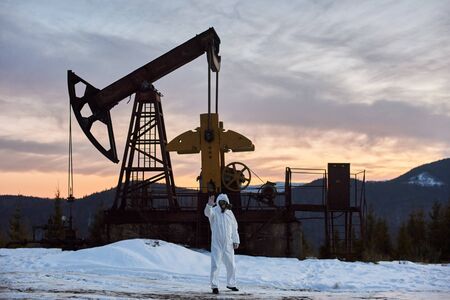 Horizontal snapshot of an oil rig in the mountainous area against purple sunset and mountain ridge, researcher in protective suit showing stop sign in front of the oil pump jackの写真素材