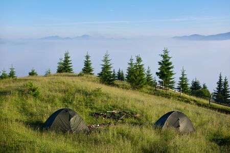 Two tents laid out on a green hill in the summer mountains in sunny morning facing incredible rising fog from a valley, concept of active lifestyleの写真素材