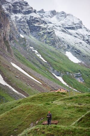 Young man hiker with trekking poles standing near wooden bench in mountain village. Beautiful view...