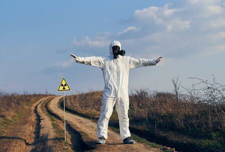 Full length of male scientist standing on the road near yellow triangle with ionizing radiation warning sign and spreading arms to sides. Research scientist wearing protective suit, gas mask.の写真素材