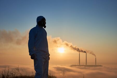 Silhouette of scientist wearing a white coverall and gas mask standing on a hill, smoking pipes of...