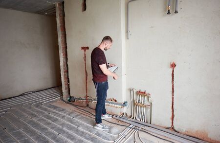 Young man in empty unfinished apartment measuring the wall with a help of measure tape, checking notes, standing on floor heating pipe system. Concept of repair and refurbishment.の写真素材