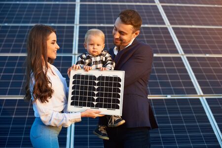 Young happy family on the background of solar panels. A man, woman and child are holding a solar panel in their hands and smiling. Solar energy concept imageの写真素材