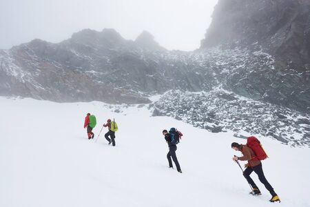 Group of brave hikers with trekking sticks and backpacks having winter hiking tour in mountains, walking through snow near big rocky hill. Concept of travelling, hiking and mountaineering.の写真素材