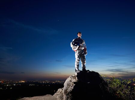 Brave astronaut standing on top of rocky hill with beautiful blue sky on background. Handsome spaceman in white space suit holding helmet. Concept of cosmonautics and space travel.の写真素材