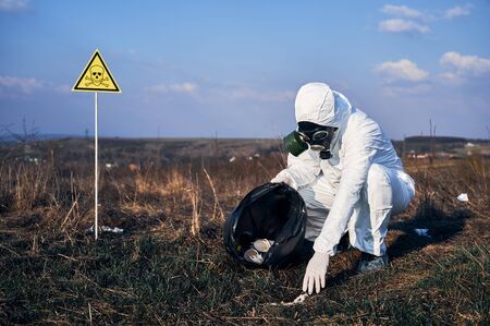 Male environmentalist in protective suit and gas mask putting trash into garbage bag with warning symbol of poisonous substances and danger. Concept of ecology, environmental pollution and hazard.の写真素材