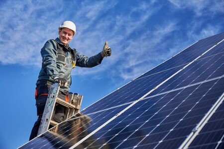 Portrait of a smiling worker, installing solar batteries, standing on ladder at solar plant against blue sky, showing thumb up. Concept of alternative energy and powerの写真素材