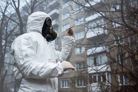 Man in gas mask and protective clothing monitoring city environment. Scientist checking tree leaves, taking piece using tweezers for analysis. Environmental disaster, smoky empty atmosphere in yard.の写真素材