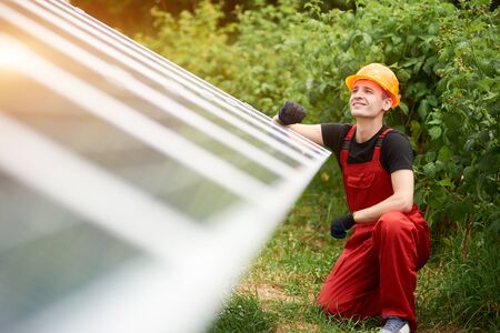 Young technician in orange uniform, safety cap near on blurry background of solar panels and...
