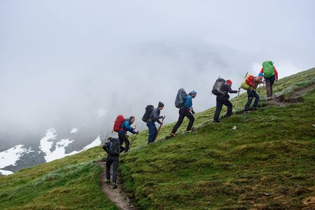 Group of male hikers with backpacks using trekking sticks while climbing grassy hill. People walking uphill in mountains with foggy cliff on background. Concept of hiking, travelling and backpacking.の写真素材