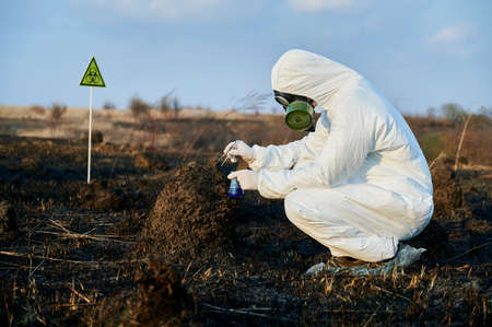 Side view of male ecologist in protective suit taking burnt grass and soil samples, holding test...
