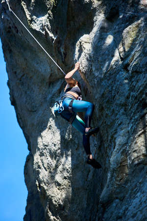 Brave female alpinist in sportswear using rope while climbing rock formation on sunny day. Young...