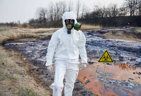 Scientist wearing white protective coverall and gas mask standing next to a biohazard sign at...