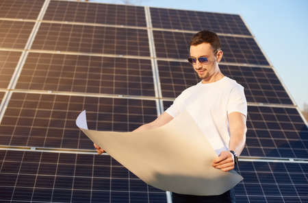 Young businessman is looking into a white paper plan standing by a big solar panel, sun is shining brightly on the face, eco-friendly conceptの写真素材
