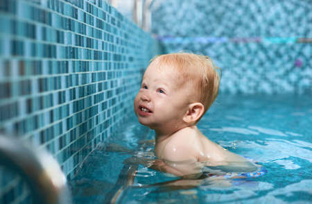Cheerful blonde little boy is trying to swim holding a silver pipe in the swimming pool. Concept of recreational sport for infantsの写真素材