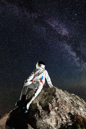 Astronaut resting on top of rocky mountain under beautiful night sky with stars. Astronaut wearing white space suit with helmet. Concept of space exploration, cosmonautics and milky way.の写真素材