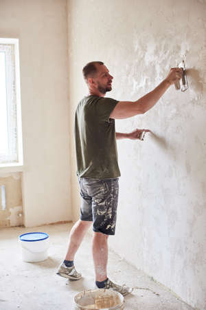 Master is applying white putty on wall and smearing by special knife in room of renovating apartment at daytime. Guy in smeared shorts and T-shirt near bucket with mixture against new windowの写真素材