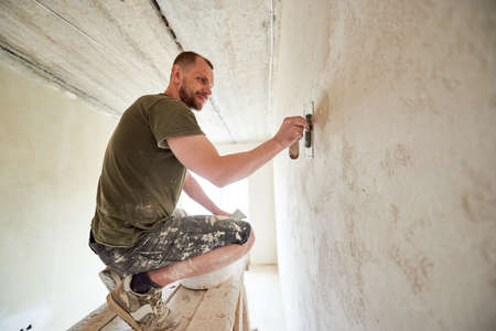 Guy at wooden stand is puttying the walls indoors small room in the daytime. Guy with beard in...