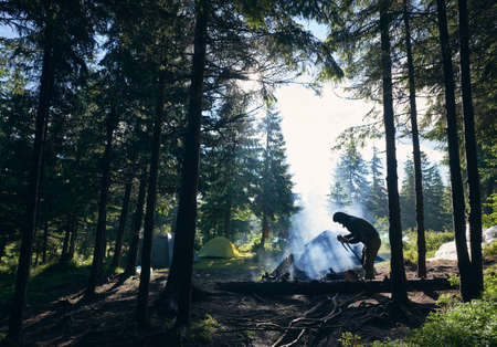 Silhouette of male tourist bending over wooden trunk, breaking branches, trying to make fire near tents. Camping, tourism in summer spuce forest, fresh air, sunny day.の写真素材