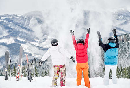 Back view of friends skiers in ski suits and helmets throwing fresh powder snow high in the air, having fun on snowy hill with beautiful mountains on background. Concept of ski resort and friendship.の写真素材