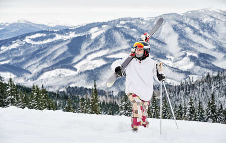 Horizontal snapshot of young skier in colorful costume walking along snowy hill, carring his skis, against amazing mountains on background. Front view, copy space, full lengthの写真素材