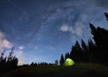 Beautiful summer night in the mountains, magical sky full of stars over high tops of spruce trees and illuminated green tent in the middle of mountain meadow. Copy space. Concept of tourism and natureの写真素材