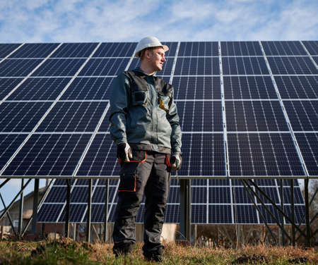 Full length of man electrician in safety helmet looking away while maintaining solar photovoltaic panel system. Concept of alternative energy and power sustainable resources.の写真素材