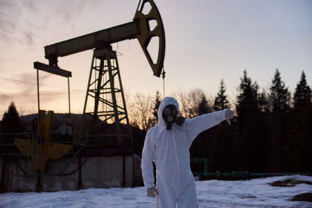 Scientist in gas mask, protective suit showing disapproval gesture while standing on territory of oil field with pump jack on background. Concept of petroleum industry and environmental pollution.の写真素材