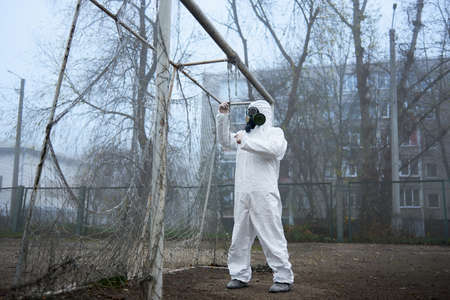 Man in gas mask and protective clothing monitoring abandoned football pitch in emty city, fog...