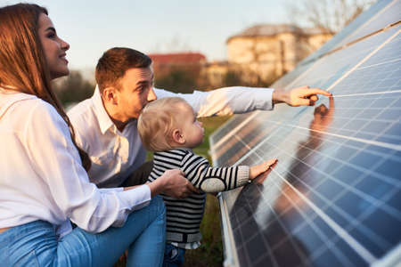 Side close-up shot of a young modern family with a little son getting acquainted with solar panel on a sunny day, green alternative energy conceptの写真素材