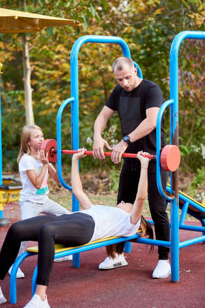 Young family is having active time at the outdoor gym, wife is lying down and lifting red barbell...
