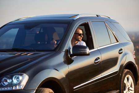 Handsome man is sitting in his black car looking out of the window wearing sunglasses, looking away, outside the city, glimpse of the setting sun, side viewの写真素材
