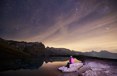Scenery at night, woman sitting on stone near lake, beautiful mountains with snow on background....