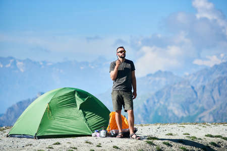 Male tourist enjoying scenery, standing and talking by radio near tent in the rocky alpine mountains. Hiker in shorts and sunglasses resting in mountains. Concept of nature, camping in Alpsの写真素材