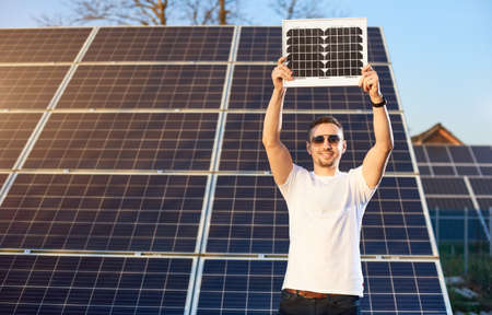 Stylish guy holds a solar panel above himself. In the background are rows of solar panels in the...