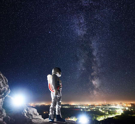 Mission specialist spaceman standing on rocky hill and looking at beautiful starry sky with milky way. Space traveler wearing white space suit and helmet. Concept of human space exploration.の写真素材
