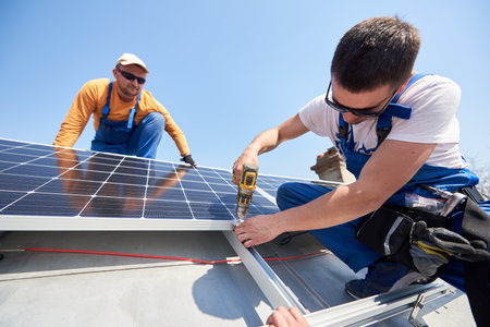 Male team workers installing solar photovoltaic panel system using screwdriver. Two electricians mounting blue solar module on roof of modern house. Alternative energy innovation concept.の写真素材