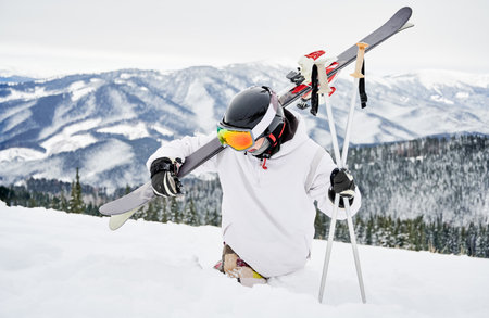Alpine skier in snowboard jacket holding skis and ski poles while trying to walk through deep white snow. Man spending vacation in mountains. Concept of winter sport activities.の写真素材