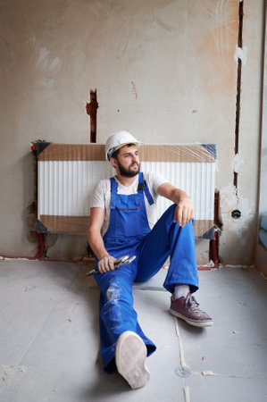 Young plumber wearing working uniform and helmet, sitting on the floor, looking away, holding wrench, resting on radiator after finishing installation works. Concept of heating system installation.の写真素材