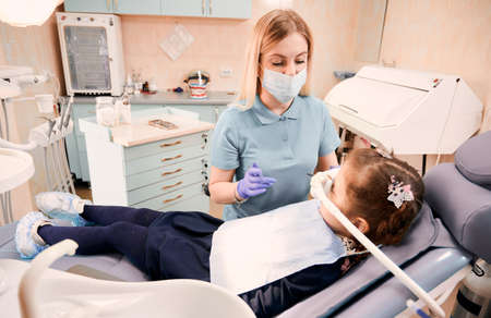 Young woman dentist in medical face mask holding dental instruments while little girl lying in dental chair with inhalation sedation. Concept of sedation dentistry.の写真素材