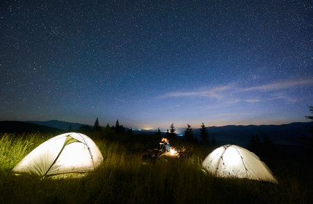 Magnificent view of night starry sky over grassy valley with hikers and illuminated camp tents. Man with his girlfriend sitting near campfire. Concept of hiking, night camping and relationships.の写真素材