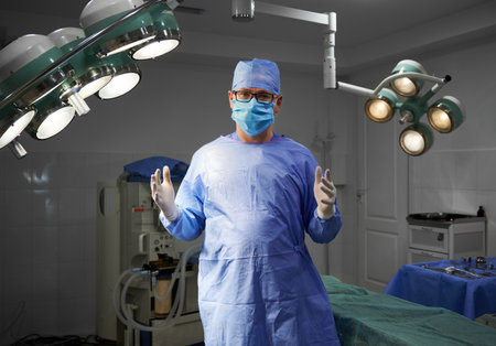 Portrait of surgeon looking at camera while standing in operating room at hospital. Man doctor wearing surgical uniform, sterile gloves, protective face mask, medical cap. Concept of medicine, surgeryの写真素材
